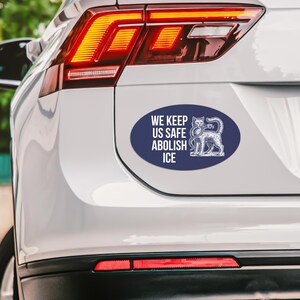 May include: An oval-shaped bumper sticker with the text "WE KEEP US SAFE ABOLISH ICE" in white on a navy blue background. A detailed illustration of a cat is on the right side of the sticker.