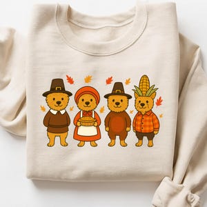 May include: Beige sweatshirt featuring a Thanksgiving-themed design. The graphic depicts four cartoon bears dressed in festive attire, including pilgrim hats, aprons, and a corn husk headdress, surrounded by autumn leaves.