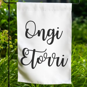 May include: White garden flag with black text that reads "Ongi Eturri", which is a Basque phrase meaning "Welcome".