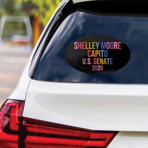 May include: An oval-shaped black car decal featuring multi-colored text that reads: SHELLEY MOORE CAPITO U.S. SENATE 2026. The sticker is applied to the rear window of a white vehicle, displaying the political candidate's name and election year.