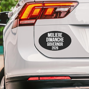 May include: An oval-shaped black bumper sticker with white text that reads MOLIERE DIMANCHE GOVERNOR 2026, applied to the rear trunk of a white vehicle featuring a red taillight assembly and a reflective red light strip along the lower bumper.