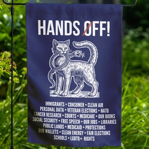 May include: A navy blue garden flag with white text and a cat illustration. The text reads "HANDS OFF!" above a cat holding a snake. Below the cat are listed political topics such as "IMMIGRANTS" and "LGBTQ+ RIGHTS".
