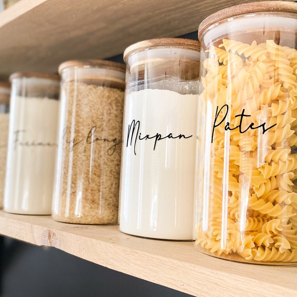 Puede incluir: Cinco tarros de vidrio con tapas de madera en un estante de madera. Los tarros están llenos de varios productos secos, incluyendo pasta, harina y arroz. Los tarros están etiquetados con etiquetas escritas a mano: "Pasta", "Mixpan", "Harina", "Largo" y "Arroz".
