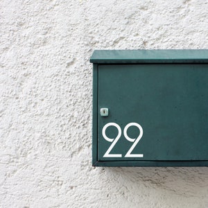 Op de afbeelding: Een groene metalen brievenbus met het nummer "22" in wit op de voorkant. De brievenbus is gemonteerd op een witte muur.