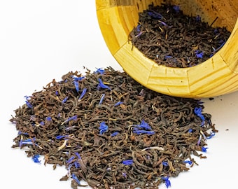 May include: A wooden bowl tipped on its side, spilling a blend of dark brown loose-leaf tea mixed with vibrant blue cornflower petals onto a surface. The tea leaves are curled and fragmented, creating a textured pile of dark brown and blue.