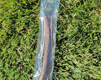 May include: A clear plastic bag containing two brown licorice sticks. The licorice sticks have a rough, textured surface. The bag is resting on a bed of green grass.