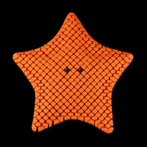 May include: An orange, star-shaped plush toy with a textured, scale-like pattern. The toy has two small, black eyes and is set against a black background. The toy is a whimsical, decorative item.