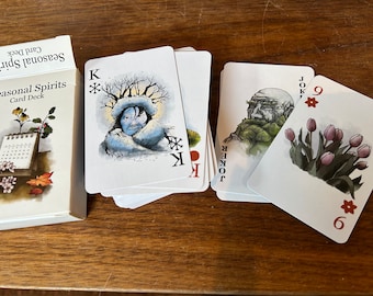 May include: A deck of playing cards with a seasonal theme. The cards feature illustrations of people and nature scenes. The box for the cards has a calendar design with a floral motif. The text on the box reads "Seasonal Spirits Card Deck".
