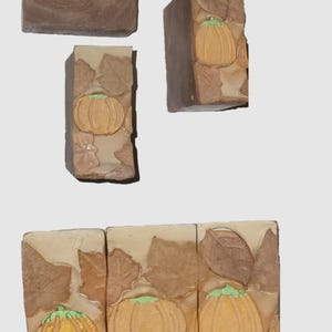 Puede incluir: Varias barras de jabón hechas a mano con temática otoñal. Los jabones son de varios tonos marrones y beige, con calabazas y hojas en relieve. Las calabazas son doradas con tallos verdes.