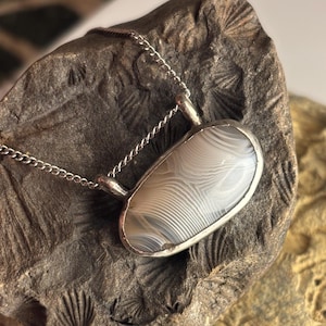May include: A silver chain necklace with a pendant featuring a white and gray agate stone. The stone has a swirling pattern and is set in a silver bezel.