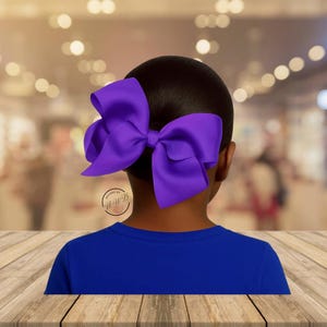 May include: A large, vibrant purple hair bow is the focal point, set against a dark background. The bow is attached to a person's dark hair, with a blue shirt visible below. The bow's design is simple and elegant, suitable for various occasions.