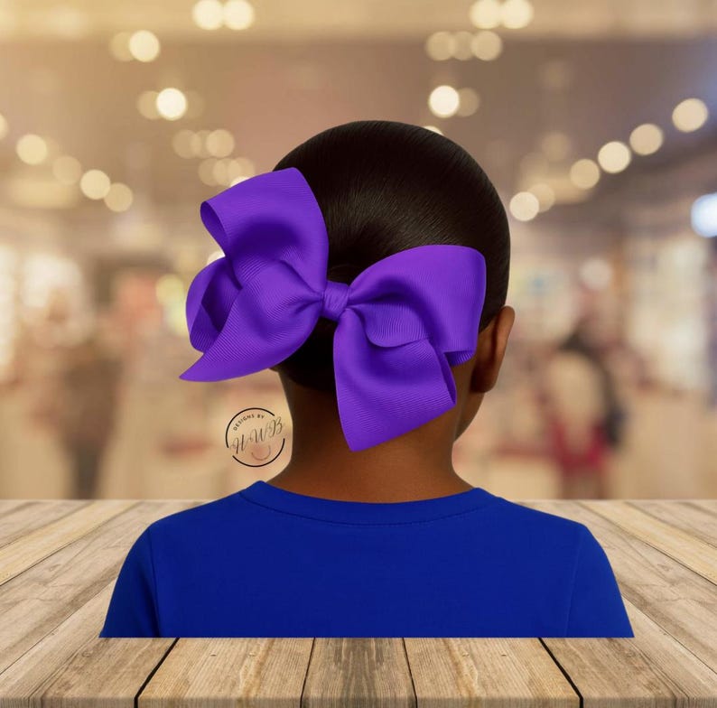 May include: A large, vibrant purple hair bow is the focal point, set against a dark background. The bow is attached to a person's dark hair, with a blue shirt visible below. The bow's design is simple and elegant, suitable for various occasions.
