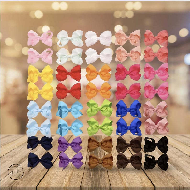 May include: A collection of colorful hair bows in various shades, including pink, white, yellow, red, orange, blue, green, brown, purple, and black. The bows are arranged in a grid pattern on a wooden surface, with a blurred background.