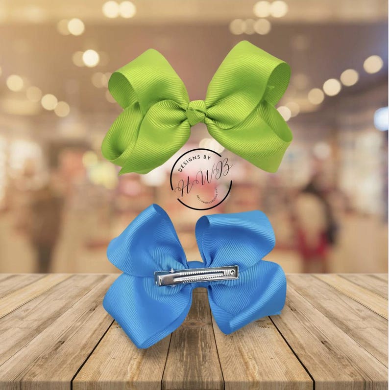 May include: Two large hair bows, one lime green and one bright blue, are displayed on a wooden surface. The bows are made of a ribbed fabric and have a classic bow shape. The blue bow has a silver clip. The image includes a logo that says "Designs by HWB".