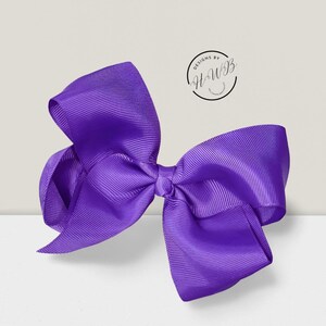 May include: A large, vibrant purple bow made of ribbed grosgrain ribbon. The bow is centered and has multiple loops and tails, creating a full, decorative appearance. The text "DESIGNS BY HWB" is in a circle in the upper right corner.