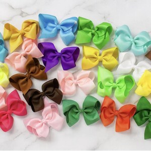 May include: A collection of colorful satin hair bows in various shades, including blue, yellow, pink, green, and brown. The bows are arranged on a white marble surface, showcasing their vibrant colors and textures. The bows are a popular accessory.
