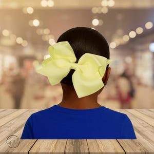 May include: A large, pale yellow hair bow is attached to the back of a person's head. The person is wearing a blue shirt. The background is blurred with bokeh lights.