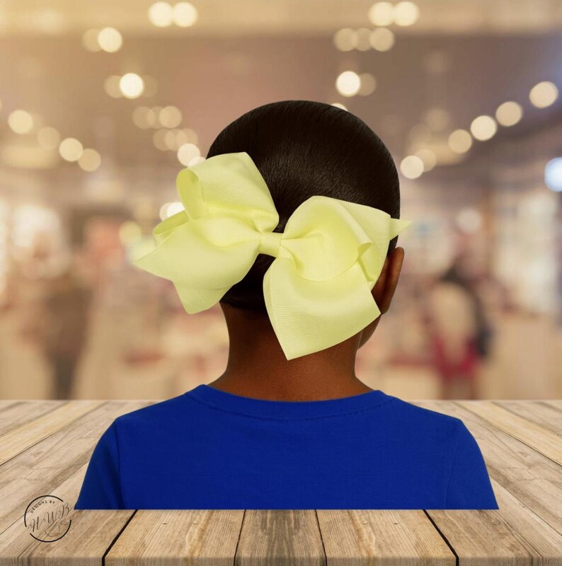 May include: A large, pale yellow hair bow is attached to the back of a person's head. The person is wearing a blue shirt. The background is blurred with bokeh lights.