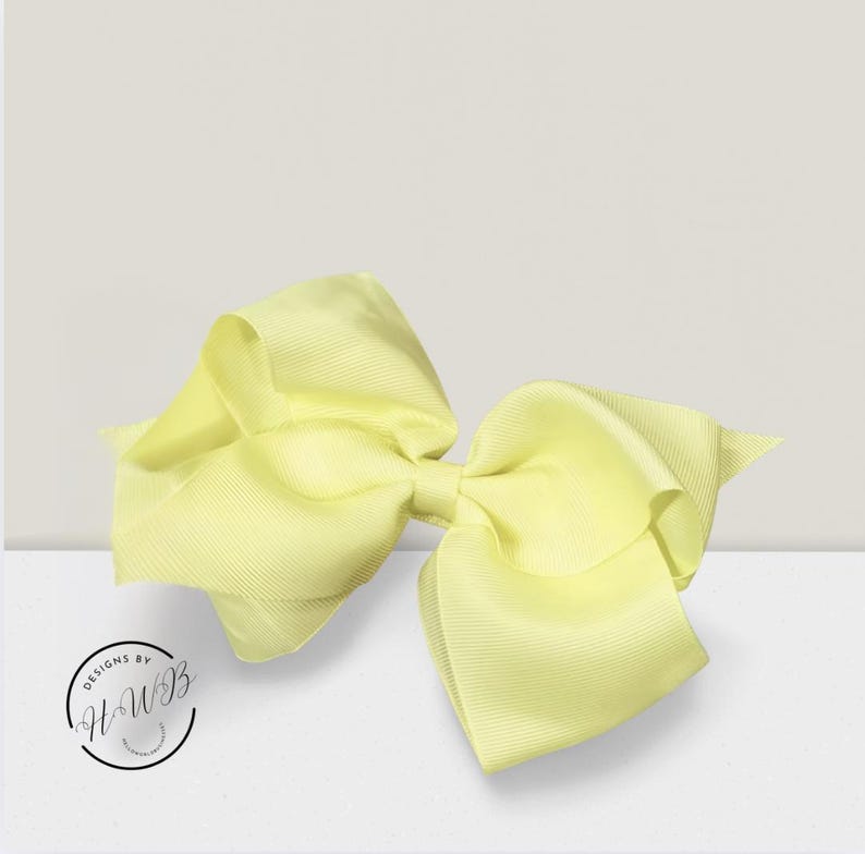 May include: A large, pale yellow hair bow made of ribbed fabric. The bow is centered and has multiple loops and tails, creating a voluminous appearance. The bow is on a white surface with a light gray background. The bow is a hair accessory.