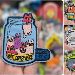 May include: A blue embroidered patch with a jar of cats and the text "Anti Depressants". There are several other patches in the background, including one with a cat in a box and the text "No Desire To Fit In", another with a cat in a litter box and the text "Trying To Get My Shit Together", and a third with a cat and the text "No Kisses Just Hisses".