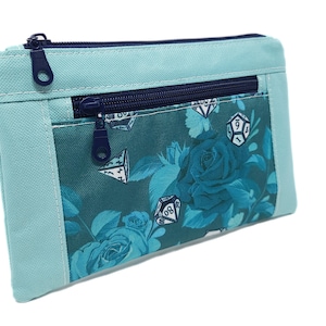May include: A teal blue zippered pouch with a floral pattern featuring blue roses and dice. The pouch has a light blue outer flap with a zipper closure.