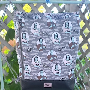 Può includere: Zaino grigio con un motivo ripetuto dei personaggi di The Munsters. Il design include immagini di Lily e Herman Munster, con il logo dello show. La parte inferiore della borsa è nera e ha una chiusura a zip e coulisse.