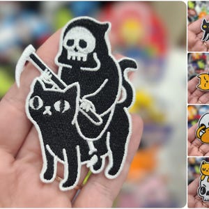May include: Black and white embroidered iron-on patch of a cat riding a skeleton on a scythe.