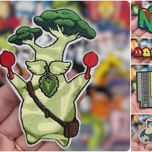 May include: Embroidered patches featuring various designs. One patch depicts a green tree-like creature holding red objects. Other patches include a colorful Nintendo 64 logo, Pac-Man ghosts, a video game cartridge with "BLOW ME" text, and a Snorlax.