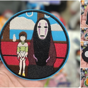 May include: A blue and red embroidered patch featuring a character from the animated film Spirited Away. The character is a black and white spirit with a face that has a white background and purple markings. The character is sitting on a red couch with a white and green character standing next to it. The patch is circular and has a blue border.