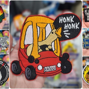 Op de afbeelding: Een verzameling geborduurde strijkpatches met verschillende ontwerpen, waaronder een gans die een auto bestuurt met de tekst "Honk Honk", een brandende vuilnisbak, een hond die op een stoel zit met de tekst "This is fine", en een patch met de tekst "Don't let the bastards get you down".