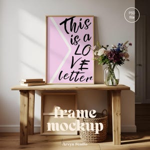 Könnte beinhalten: Ein gerahmtes Poster mit dem Text "This is a LOVE letter" auf rosa Hintergrund mit einem weißen diagonalen Streifen. Das Poster wird auf einem Holztisch mit Büchern, einer Blumenvase und einem Weidenkorb ausgestellt. Die Worte "frame mockup" sind ebenfalls sichtbar.