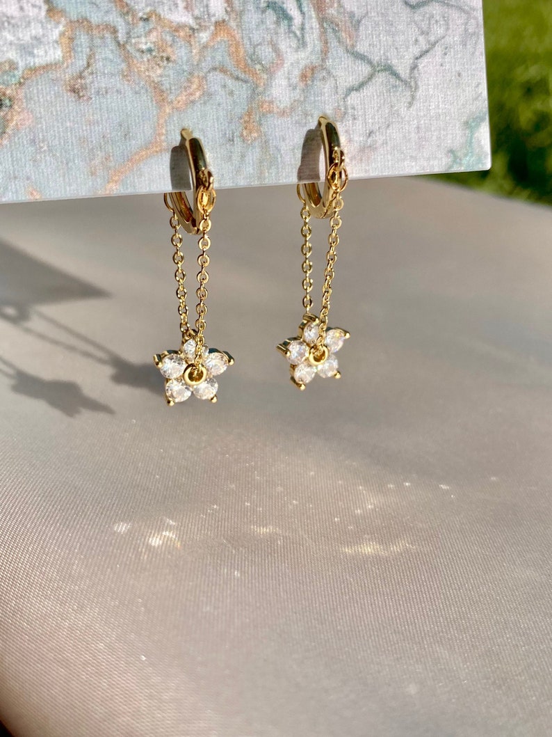 May include: Gold hoop earrings with a delicate chain and a small flower charm with clear cubic zirconia stones.