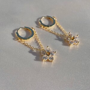May include: A pair of gold hoop earrings with a delicate chain and a small flower charm with clear cubic zirconia stones.