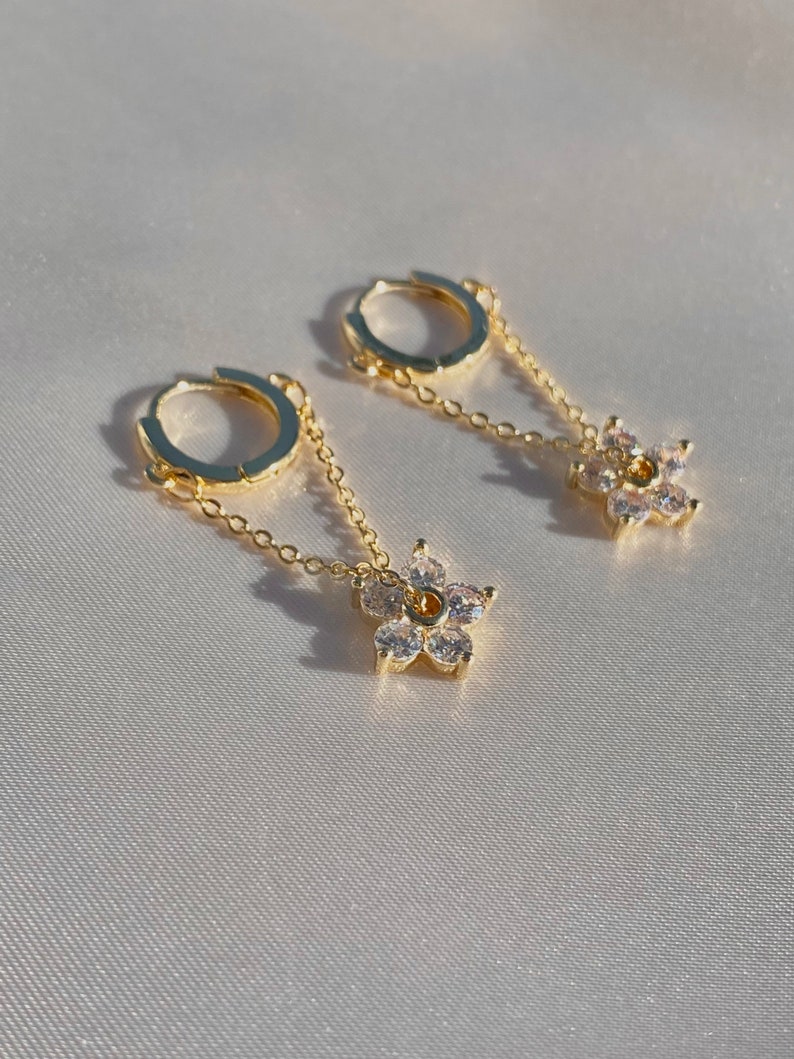May include: A pair of gold hoop earrings with a delicate chain and a small flower charm with clear cubic zirconia stones.