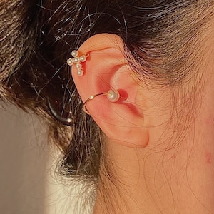 May include: A pair of gold ear cuffs with pearl accents. The ear cuff on the top is a cross shape with small pearls, while the ear cuff on the bottom is a simple hoop with a single pearl.
