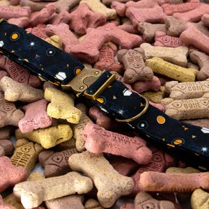May include: A black dog collar featuring a space-themed pattern with orange planets, white stars, and geometric shapes. The collar includes a metallic gold-toned buckle and a matching metal D-ring for leash attachment, resting on a pile of bone-shaped treats.