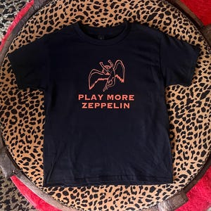 Könnte beinhalten: Schwarzes T-Shirt mit einer orangefarbenen Grafik einer geflügelten Figur und dem Text "PLAY MORE ZEPPELIN".