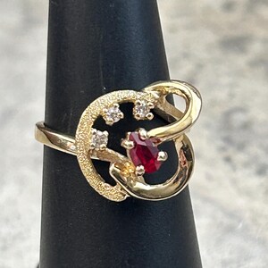 May include: A gold ring with a red gemstone and smaller clear gemstones. The ring has a unique, abstract design.