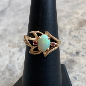 May include: A gold ring with an oval opal gemstone and two small red gemstones. The ring has a unique, abstract design.