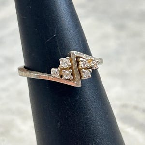 May include: A silver ring with a geometric design. The ring features a triangular shape with several small, clear gemstones clustered within the triangle and along the band. The ring is displayed on a black ring holder.