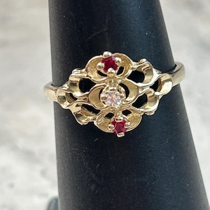 May include: A gold-toned ring with an ornate floral design. The ring features a central clear stone and two red stones, set within an openwork filigree pattern. The ring is displayed on a black stand.