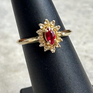 May include: A gold ring featuring a central oval-cut ruby gemstone surrounded by a halo of small diamonds. The ring's design includes a sunburst-style setting, and it is displayed on a black ring holder.