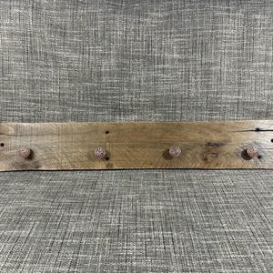 Puede incluir: Una estantería de madera rústica con cuatro pomos decorativos. La estantería está hecha de madera reciclada y tiene un acabado natural. Los pomos son de metal y tienen un acabado dorado rosa.