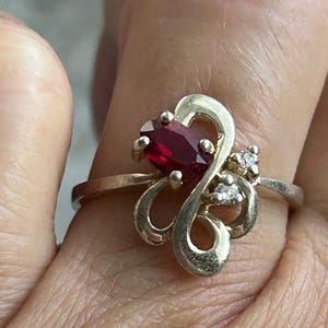 May include: A silver ring featuring a large, oval-cut, deep red gemstone as the focal point. The ring has a decorative, swirling design with small accent diamonds. The ring is worn on a finger.