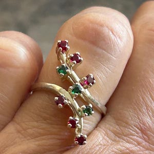 May include: A silver ring with a floral design. The ring features small, round red and green gemstones set along a curving branch-like structure. The ring is worn on a finger.