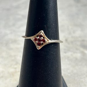 May include: A silver ring featuring a geometric design with a cluster of small, round, deep red gemstones. The ring's band is thin, and the gemstones are set within a diamond-shaped frame. The ring is displayed on a black stand.