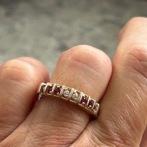 May include: A gold band ring featuring a row of alternating gemstones set in a channel style. The setting includes four round red rubies and two round clear diamonds positioned in the center of the row, all held within a polished gold metal frame.