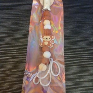 Op de afbeelding: Een fopspeenclip voor baby's met een apenontwerp. De clip heeft een witte plastic clip aan de bovenkant, gevolgd door bruine en witte kralen en een bruine apenbedel. Verpakt in een doorzichtige, iriserende plastic zak.