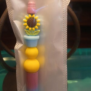 Könnte beinhalten: Ein dekorativer Stift mit Sonnenblumen-Design. Der Stift hat einen rosa Aufsatz, eine gelbe Sonnenblume, zwei gelbe Kugeln und einen blau-rosa-gelben Farbverlauf. Der Stift ist in einer transparenten Plastikhülle verpackt.