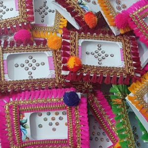 May include: A collection of rectangular cards featuring decorative bindi stickers. Each card is framed with gold metallic trim and colorful fabric fringe, accented by a single pom-pom. The stickers are arranged in various patterns.